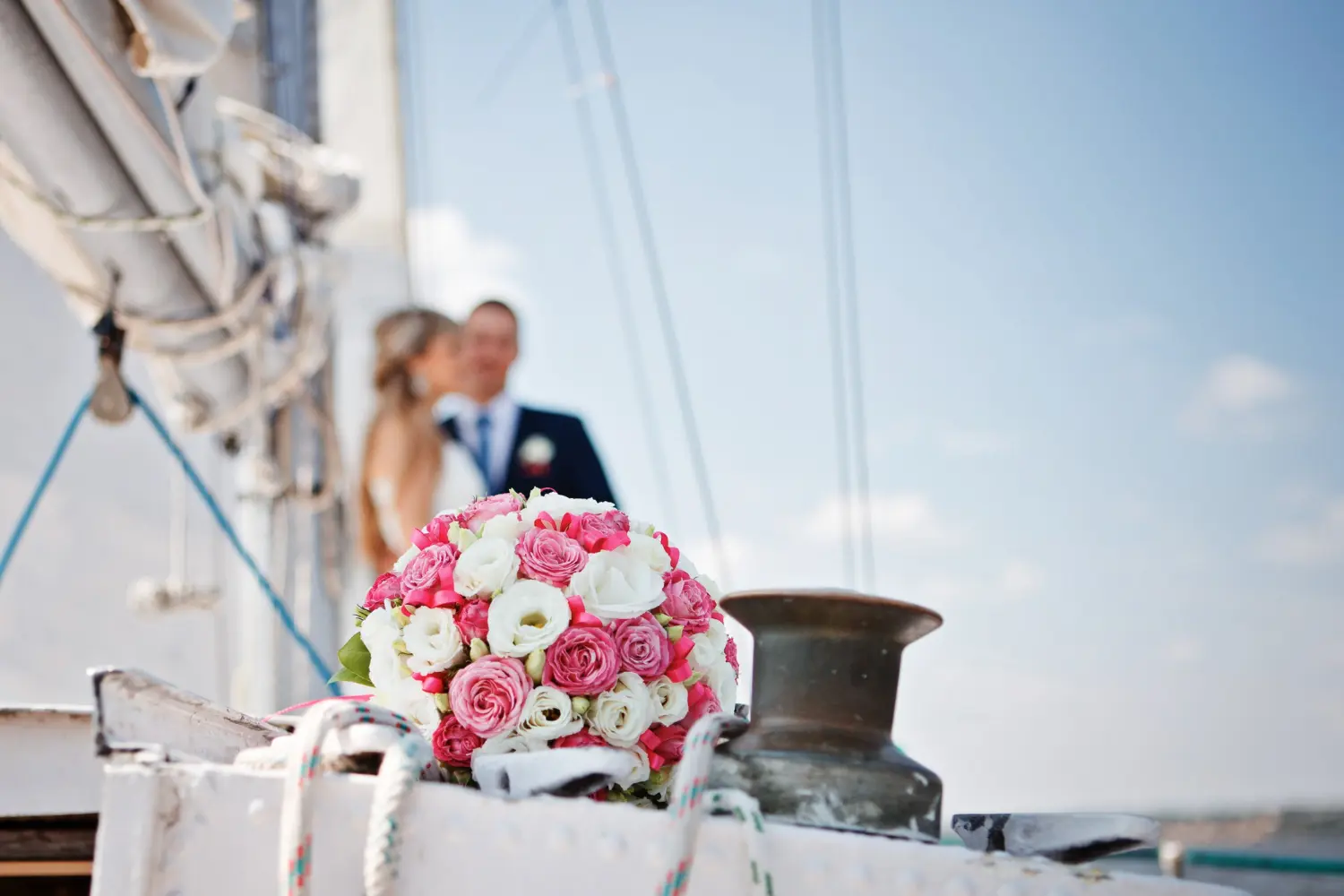 Close-up of a bridal bouquet on a yacht deck with a wedding couple blurred in the background.